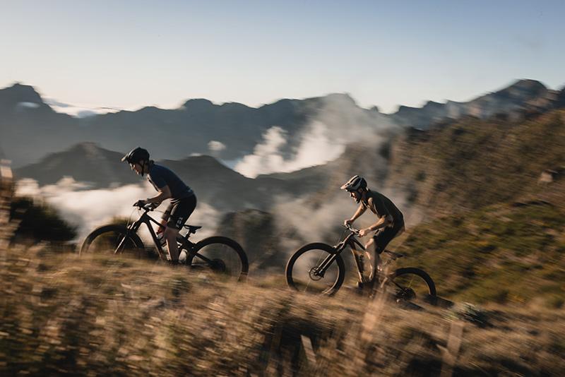 Two mountain bikers riding Trek bicycles along a rugged trail with a mountainous landscape in the background
